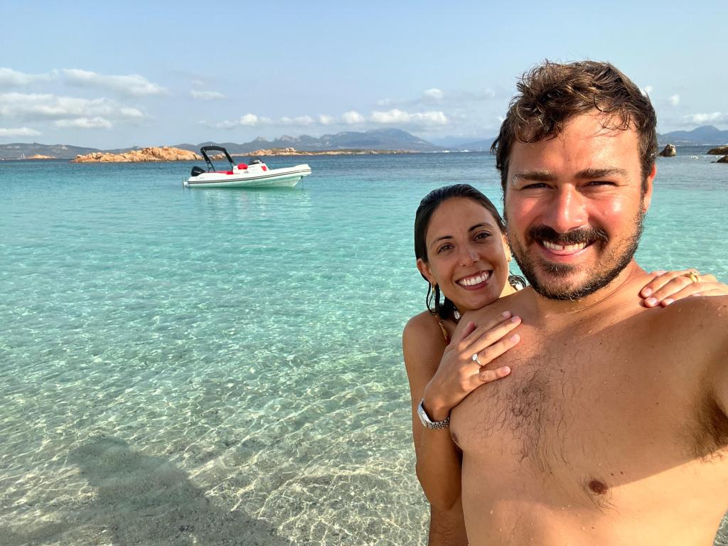 A man and woman stand in shallow water, posing for a selfie, with a boat floating in the background. The man, shirtless, h...